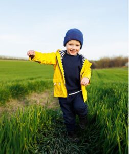 A cute little three year old boy in a yellow jacket and cap is running on a green field. A smiling child runs on green grass